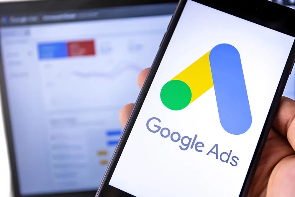 Hand holding a smartphone showing the Google Ads logo, with a blurred laptop displaying an ads dashboard behind it.