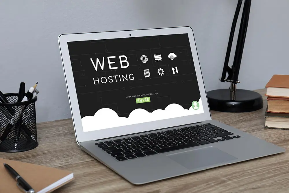 Laptop on a wooden desk showing a dark "Web Hosting" page with an Enter button, beside a desk lamp, a pen cup and books.