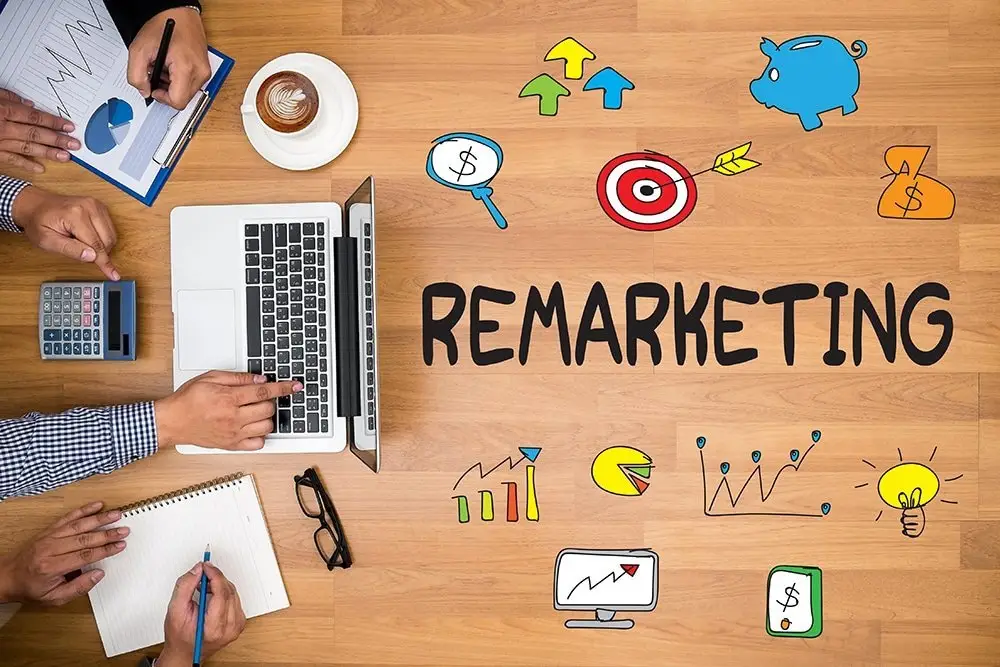 Overhead view of a wooden desk with hands at a laptop, clipboard charts and a calculator, beside doodled "REMARKETING" lettering, a target and charts