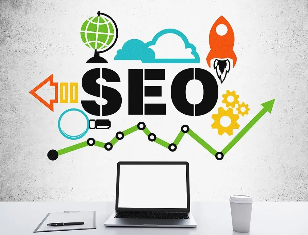 Laptop, clipboard and coffee cup on a white desk before a concrete wall painted with "SEO", a rocket, globe, gears and a rising green line chart