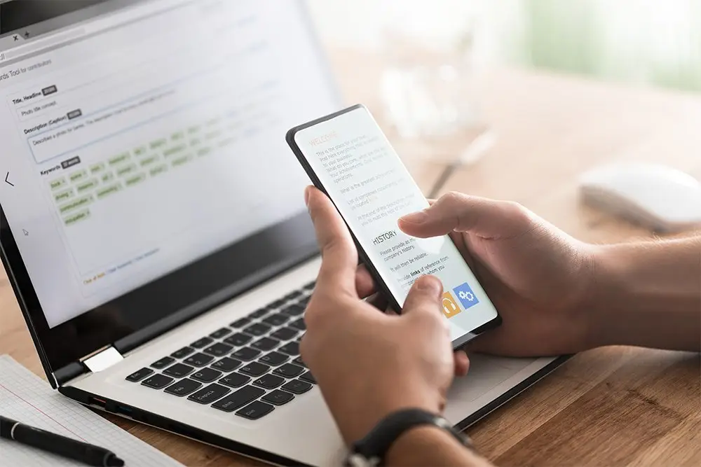 Close-up of hands holding a smartphone showing a text-heavy page, in front of an open laptop displaying a keyword entry form on a wooden desk