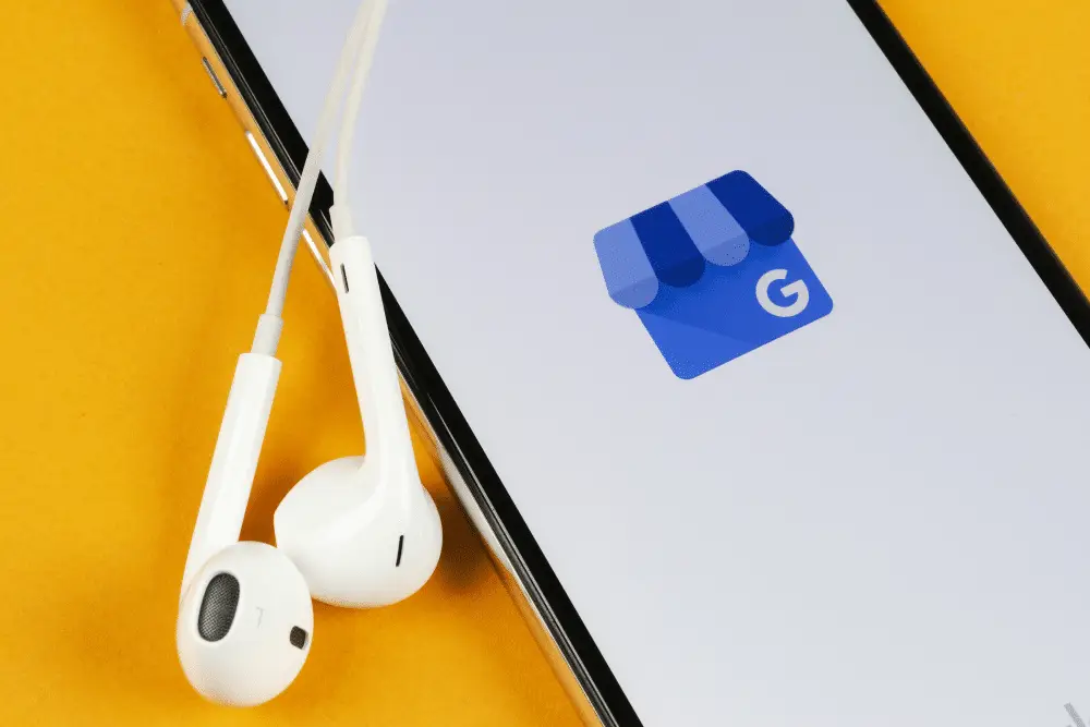 Smartphone on a yellow background showing the blue Google My Business storefront logo, with white wired earbuds beside it.
