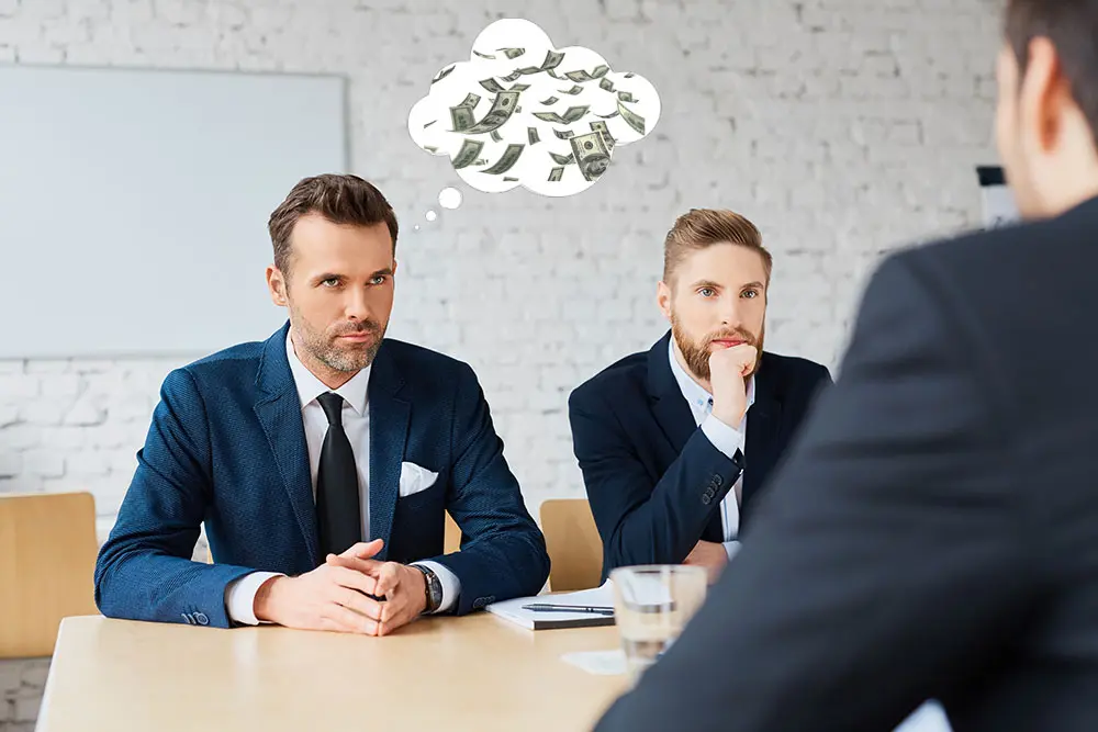 Two men in blue suits at a conference table facing a third person, with a cartoon thought bubble full of dollar bills floating above them