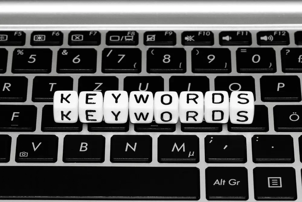 Black-and-white close-up of white letter dice spelling "Keywords" twice, resting on a laptop keyboard