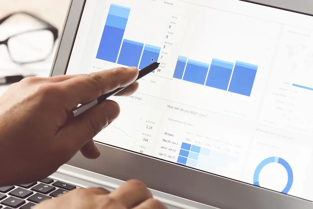 Close-up of a hand pointing a stylus at a laptop analytics dashboard with blue bar charts, a Referrals panel and a user retention table.