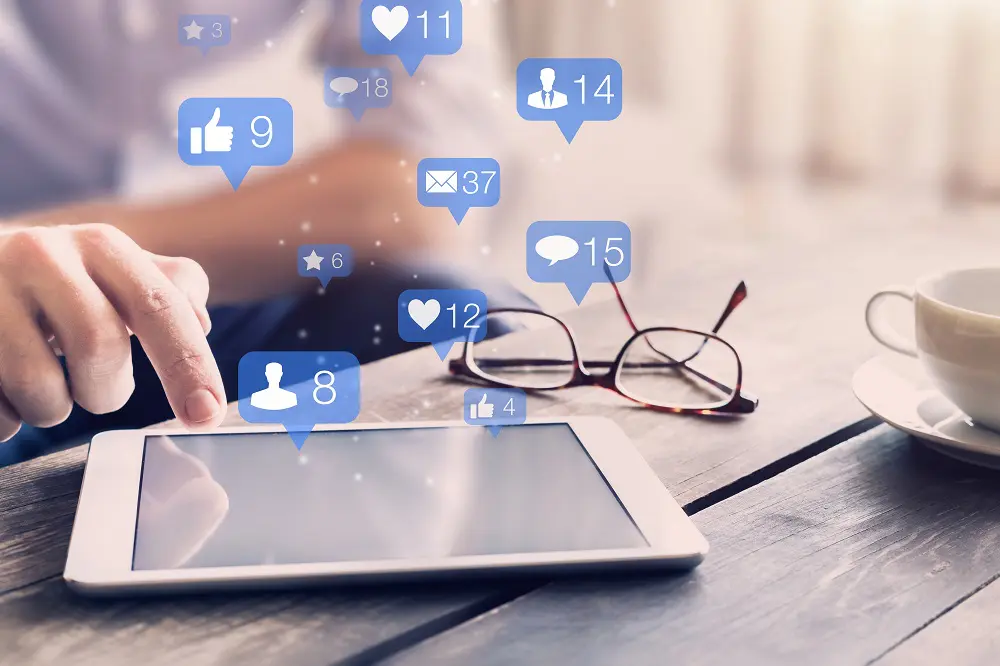 Person tapping a white tablet on a wooden table beside glasses and a coffee cup, with blue like, heart and comment notification bubbles floating above