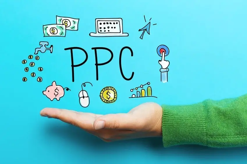 Open palm in a green sweater against bright blue, holding doodled "PPC" lettering with banknotes, a laptop, piggy bank, coins and a click cursor