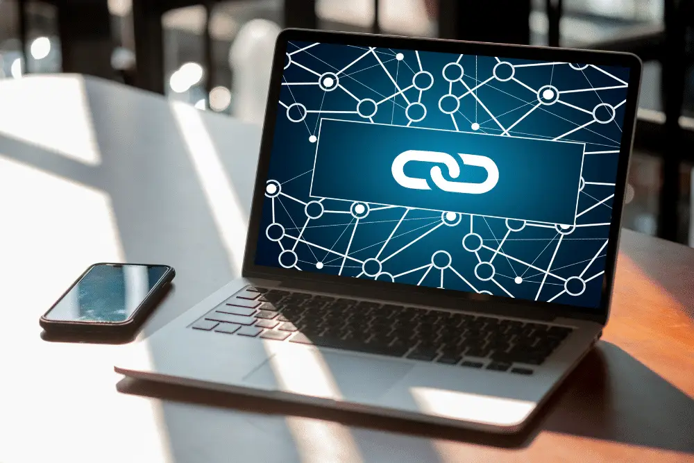 Laptop open on a sunlit white desk next to a phone, its screen showing a white chain-link icon over a dark blue network of connected nodes